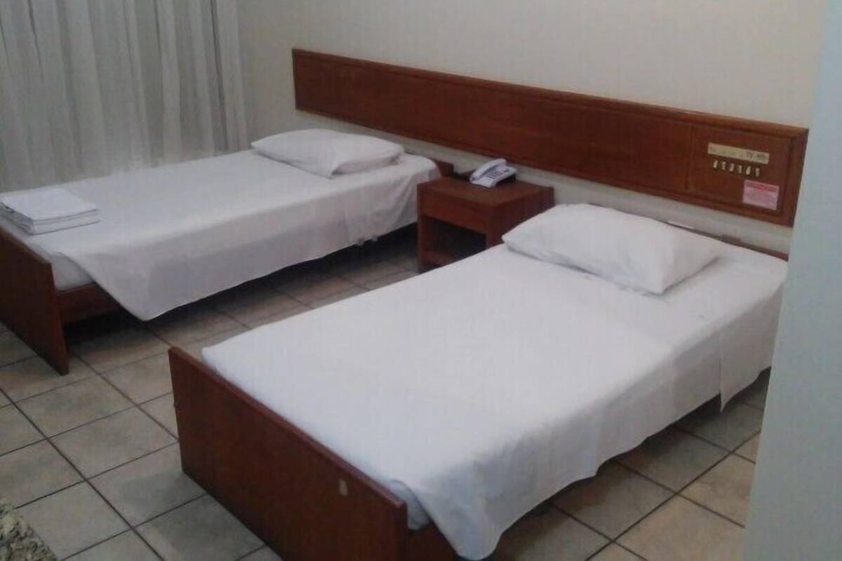A hotel room with two single beds, a wooden headboard, a bedside table with a phone, and a curtain.