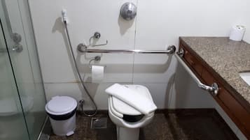 A bathroom with a toilet, a bidet, a handrail, a trash bin, and a countertop with a sink.