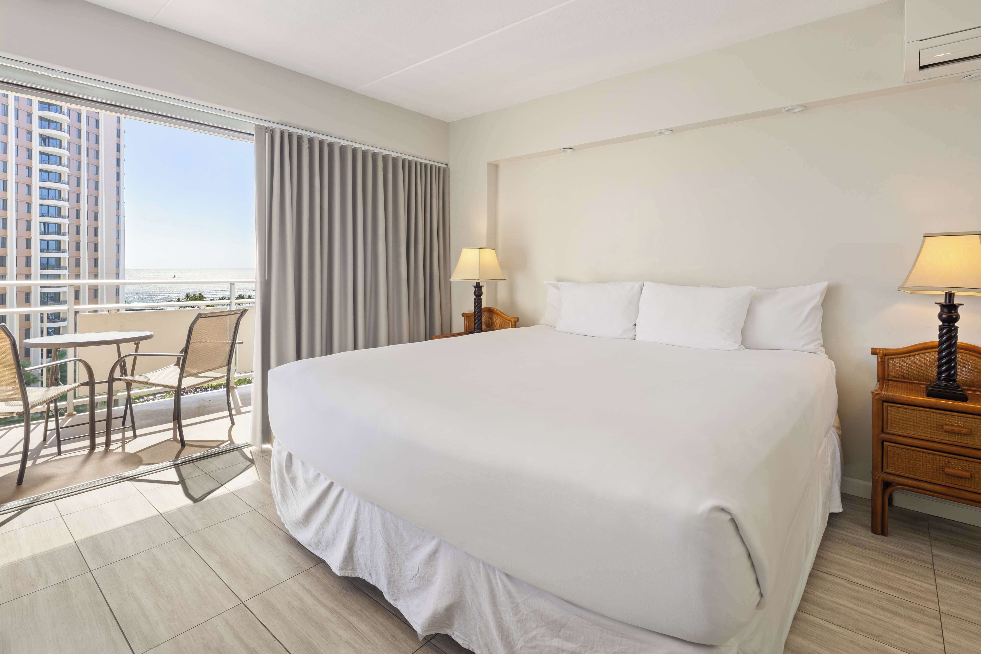 A hotel room with a large bed, two bedside tables with lamps, a small balcony with a table and chairs, and a view of a tall building and the sea.