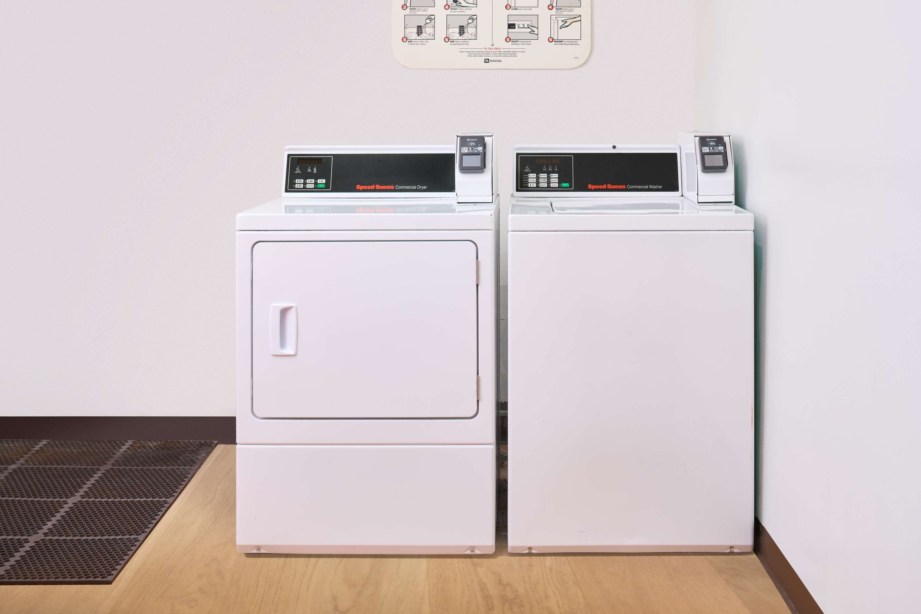 Deux sèche-linge commerciaux blancs sont placés côte à côte dans une pièce dont le sol est en bois et sur un mur est accroché un manuel d’instructions.