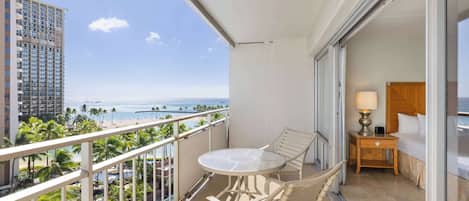 A balcony with a table and chairs, a bed with a nightstand, and a view of the ocean and palm trees.