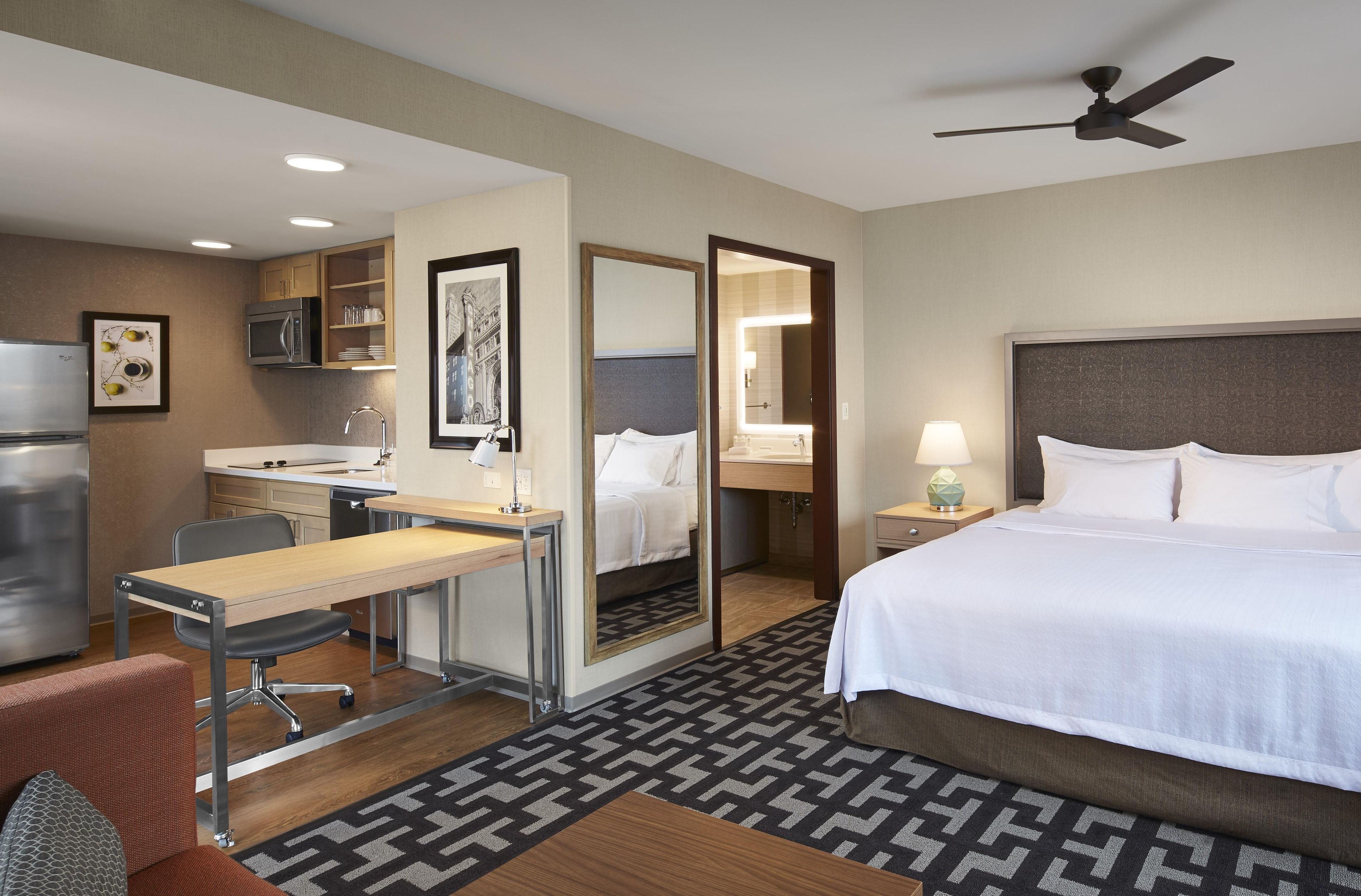 A hotel room with a bed, desk, chair, kitchenette, and a bathroom visible through an open door.