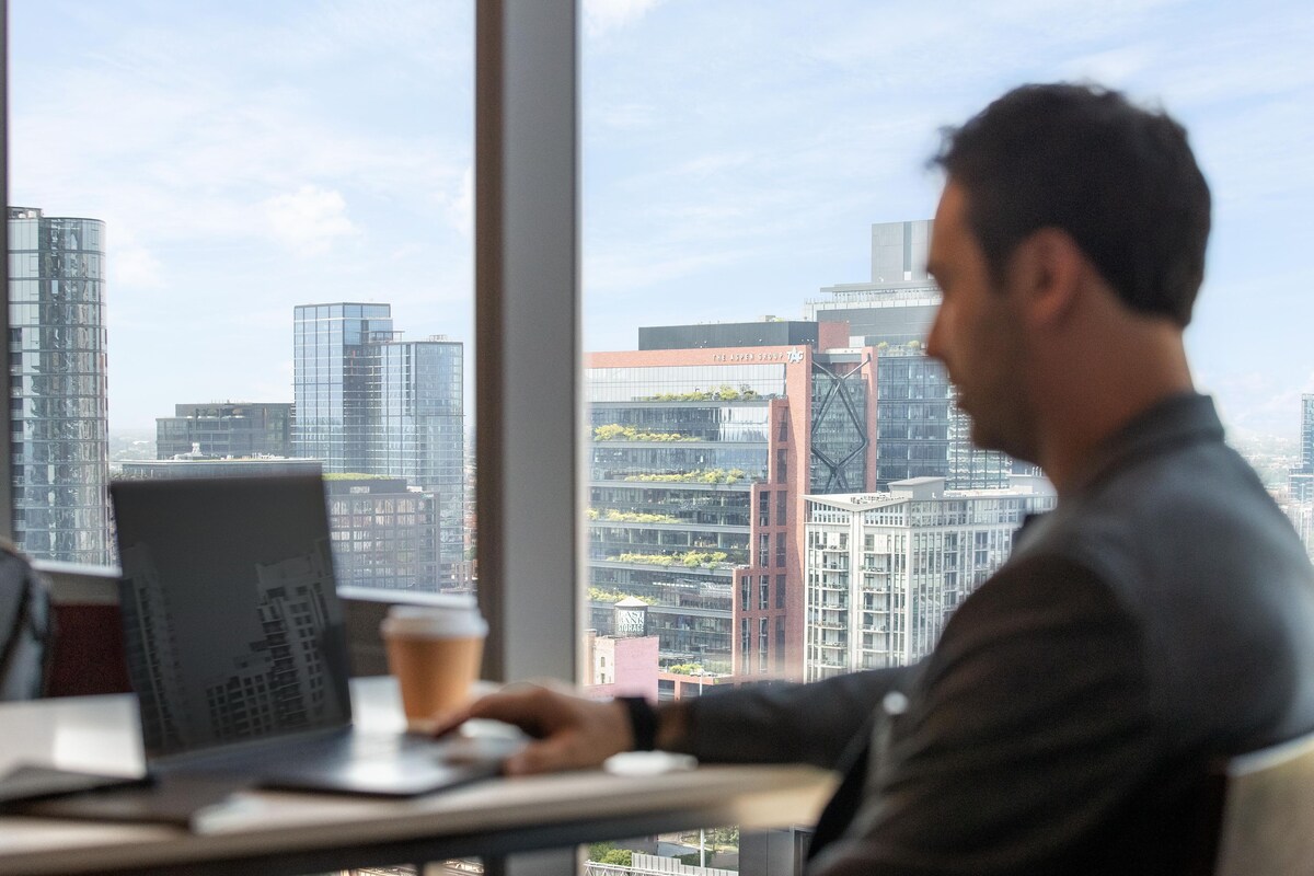 Une personne assise à un bureau, devant un ordinateur portable et une tasse de café, avec vue sur un paysage urbain composé d’immeubles modernes.