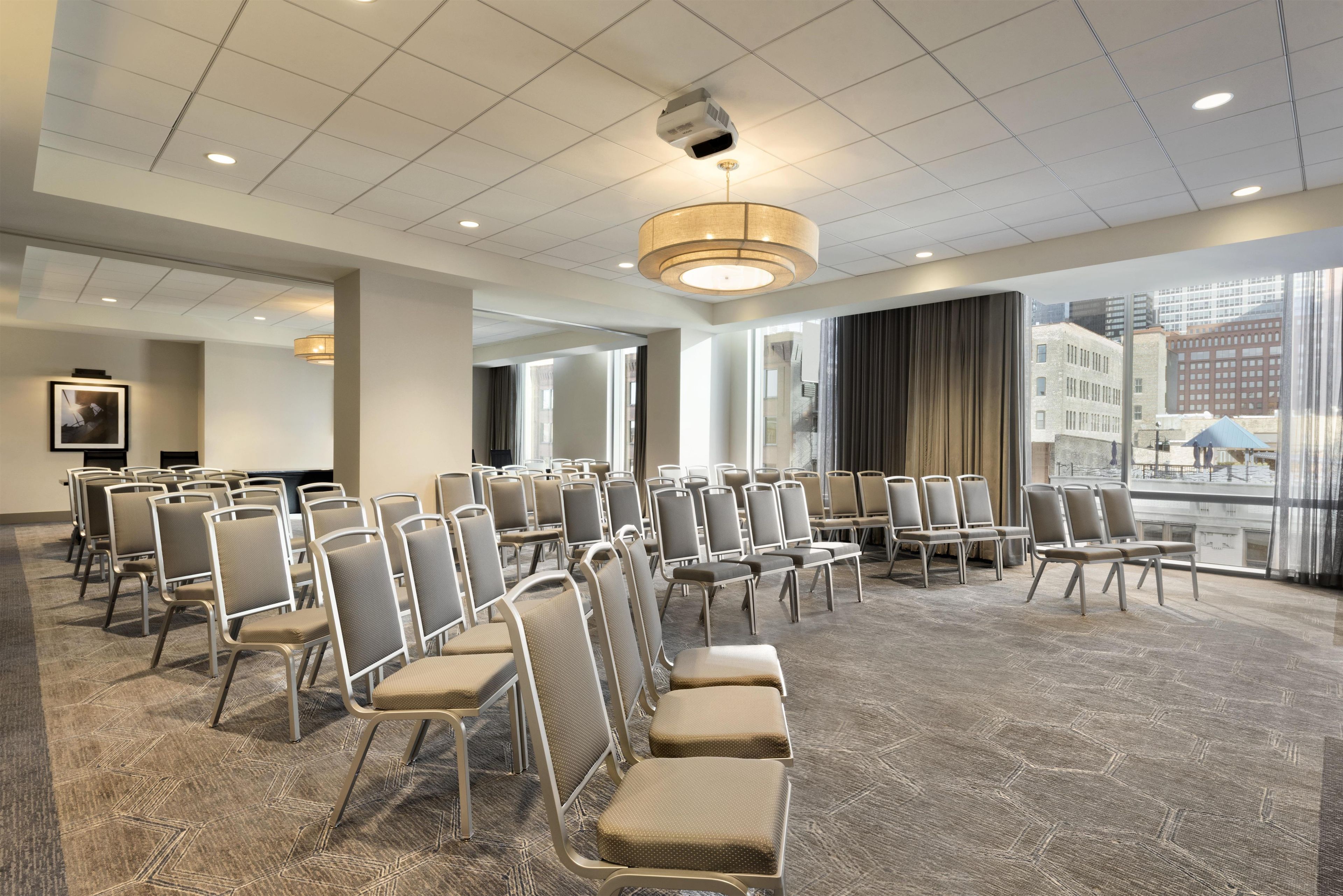 Een congreszaal met rijen stoelen, een groot raam met uitzicht op gebouwen, en een plafond met inbouwverlichting.