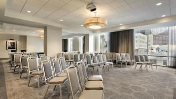 Een congreszaal met rijen stoelen, een groot raam met uitzicht op gebouwen, en een plafond met inbouwverlichting.
