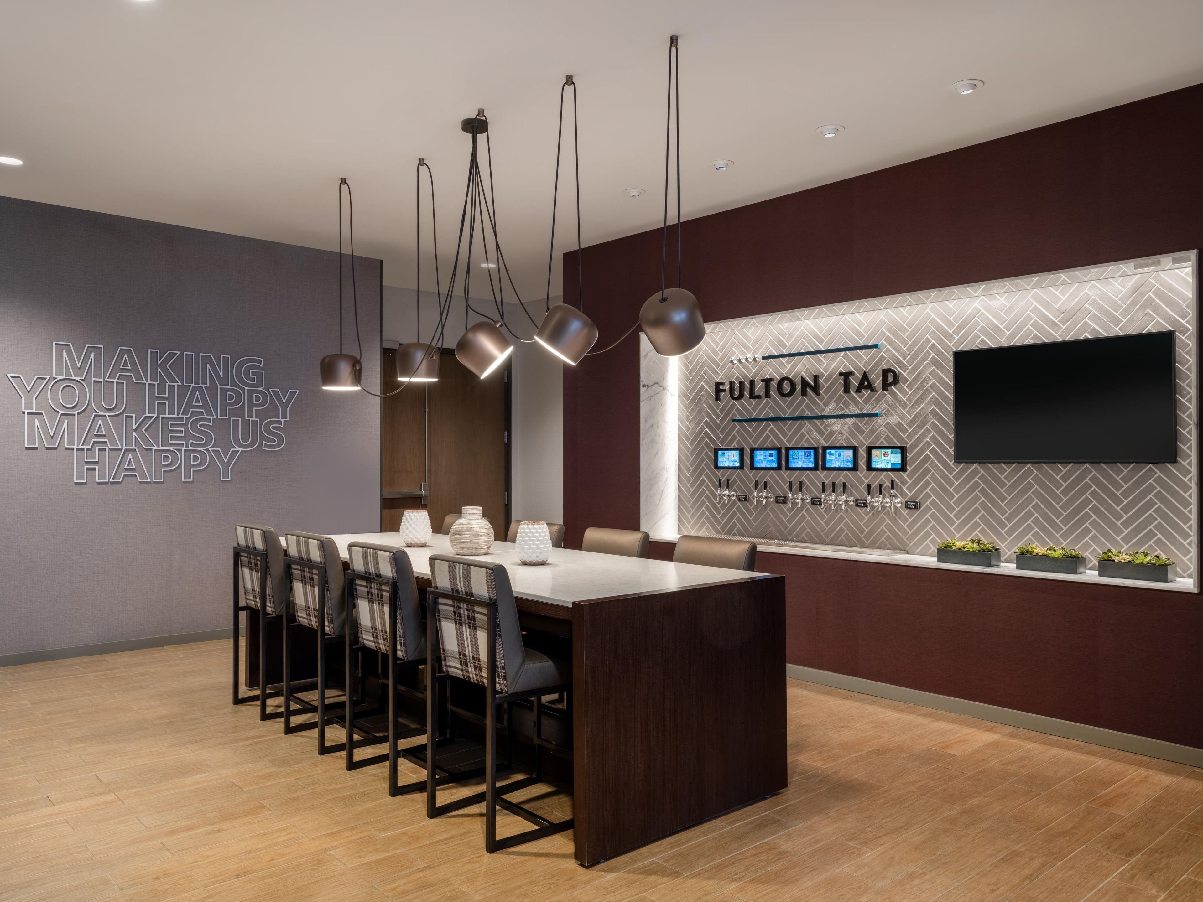 A modern dining area with a central island, multiple chairs, pendant lighting, and a wall-mounted TV.