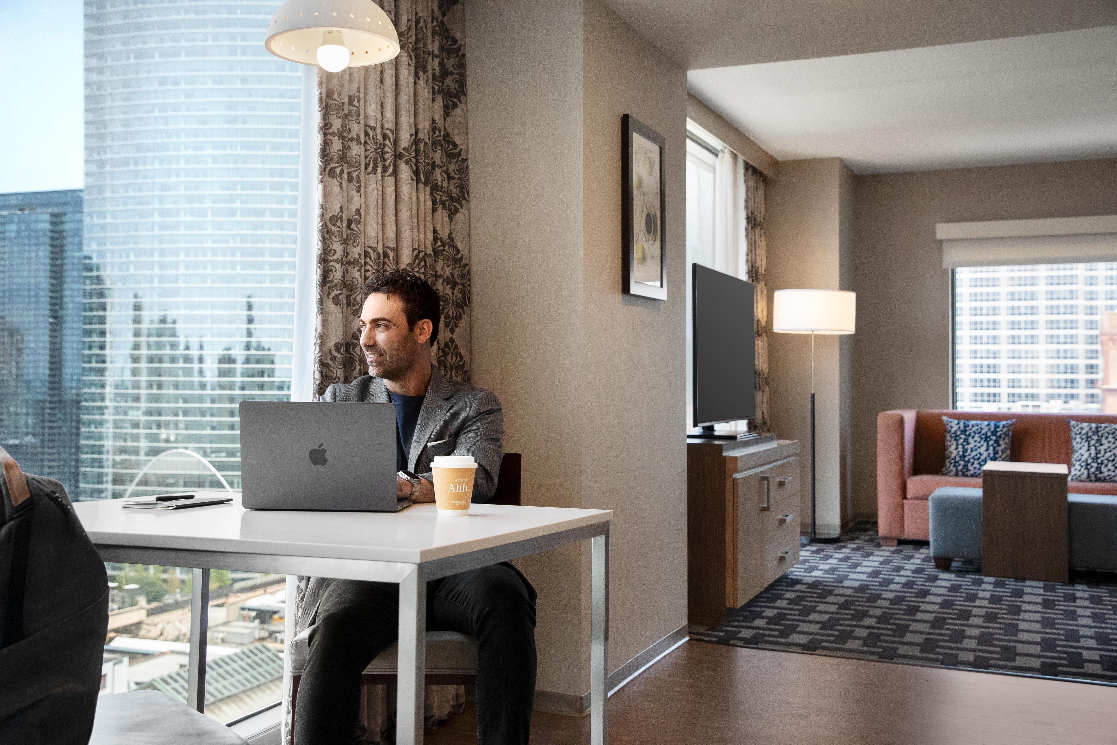 A modern hotel room with a desk, laptop, coffee cup, and a view of the cityscape.
