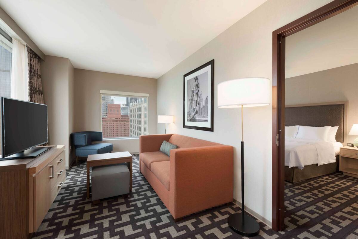 Egy szállodai szoba, amelyben van egy kanapé, egy televízió, egy íróasztal, egy lámpa és egy ágy.