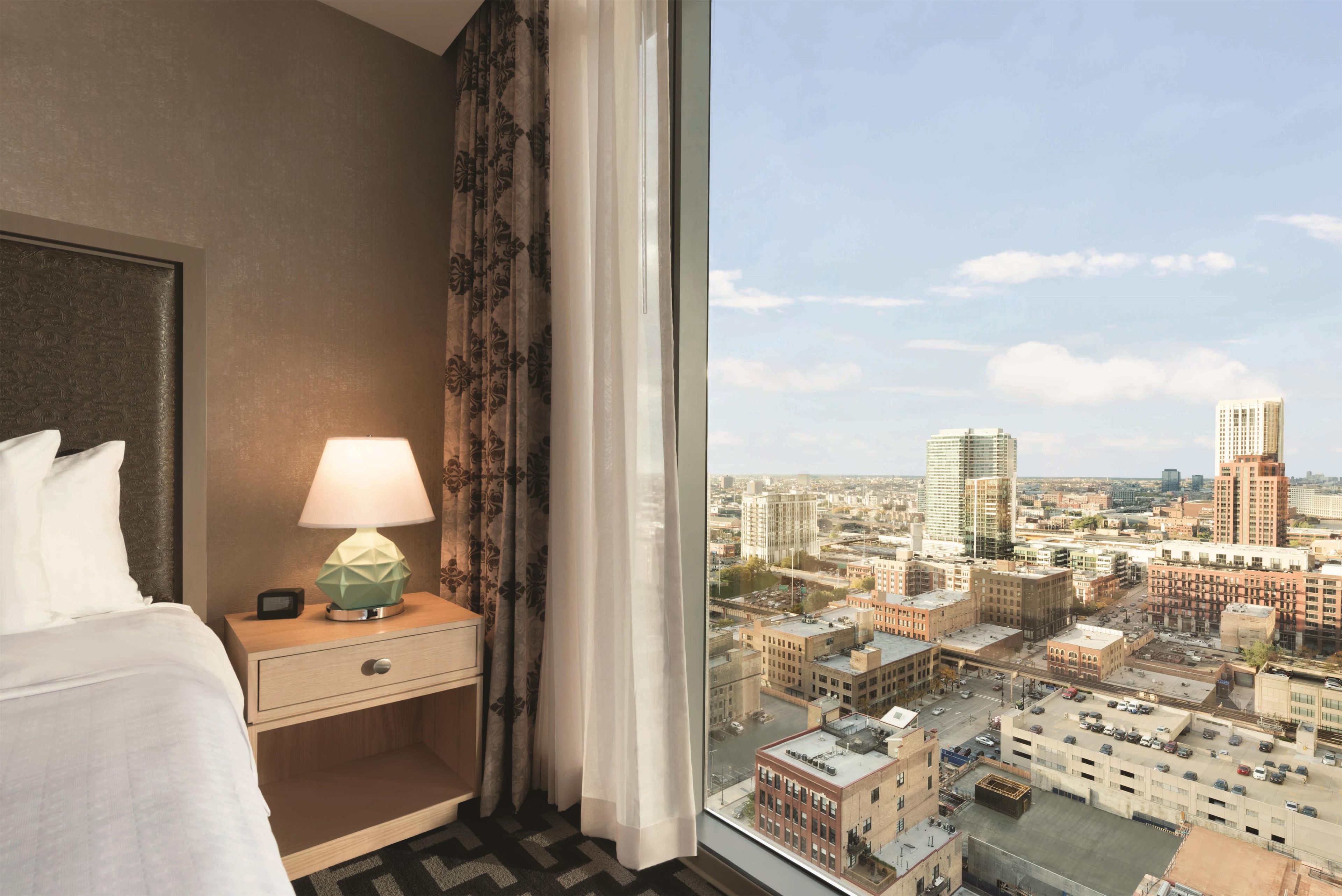 Une chambre d’hôtel avec un lit, une table de chevet avec une lampe et une fenêtre donnant sur un paysage urbain.