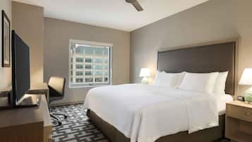 A hotel room with a bed, a desk, a chair, a television, a window with a view of buildings, and a ceiling fan.