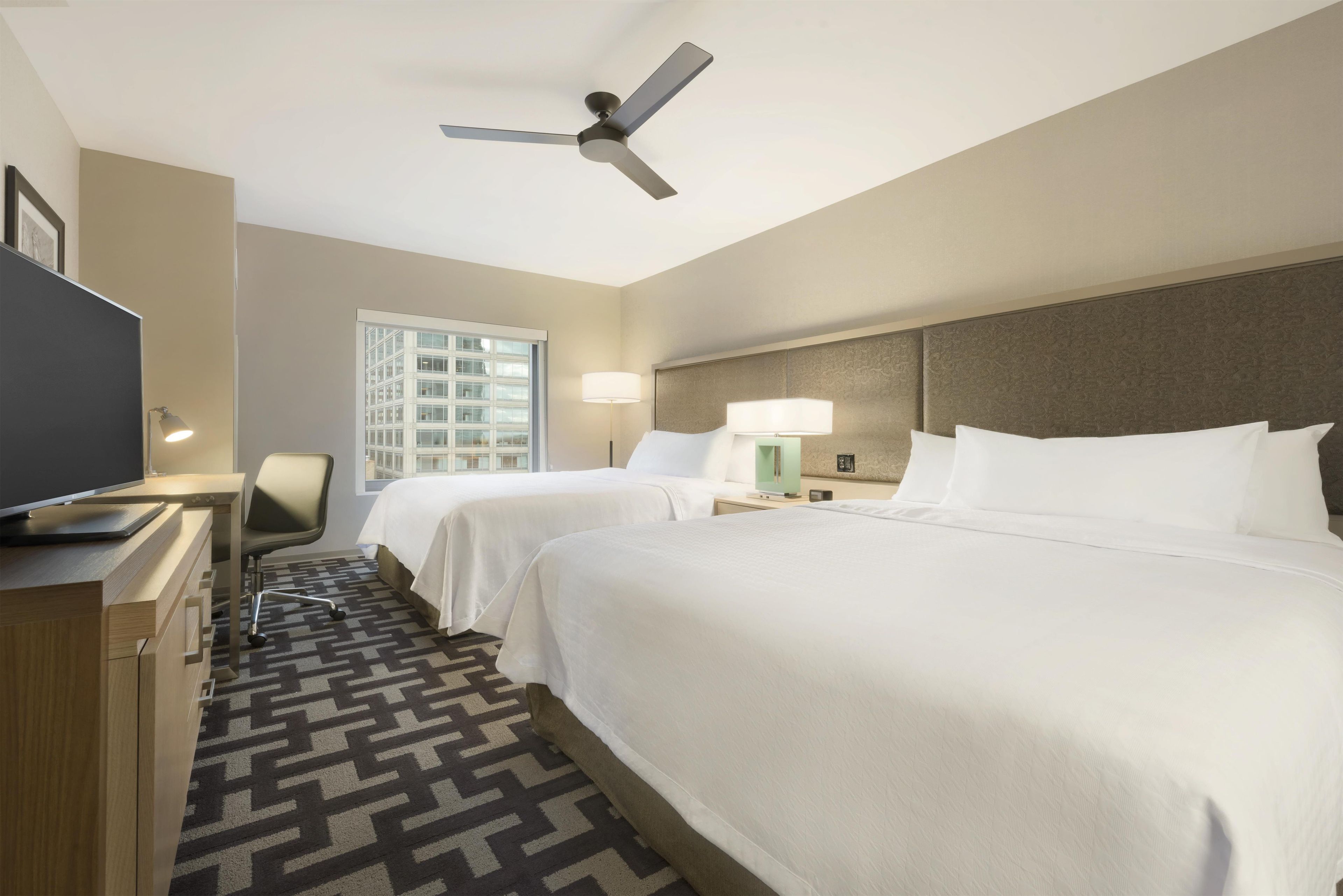 A hotel room with two beds, a desk with a flat-screen TV, a ceiling fan, and a window with a view of a building.