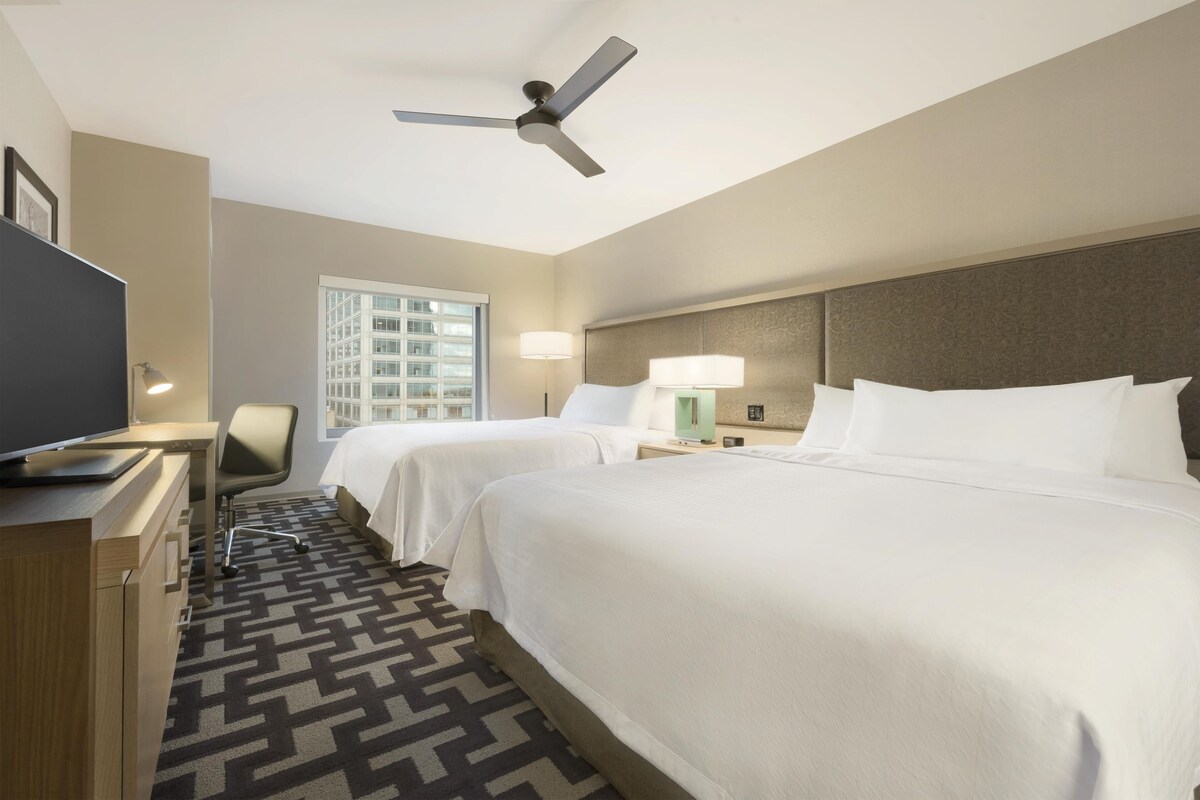 Habitación de hotel con dos camas, un escritorio, un televisor de pantalla plana, un ventilador de techo y una ventana con vistas a un edificio.