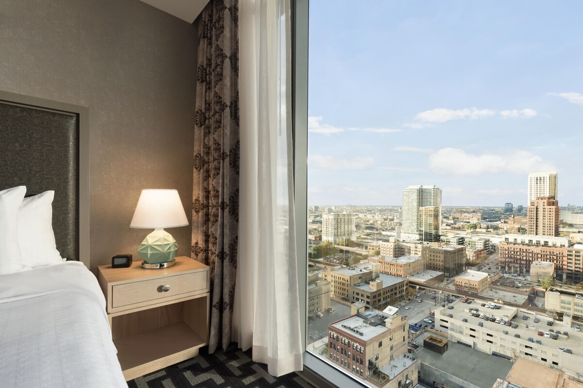 Una habitación de hotel con cama, una mesita de noche con lámpara y ventana con vista a un paisaje urbano.