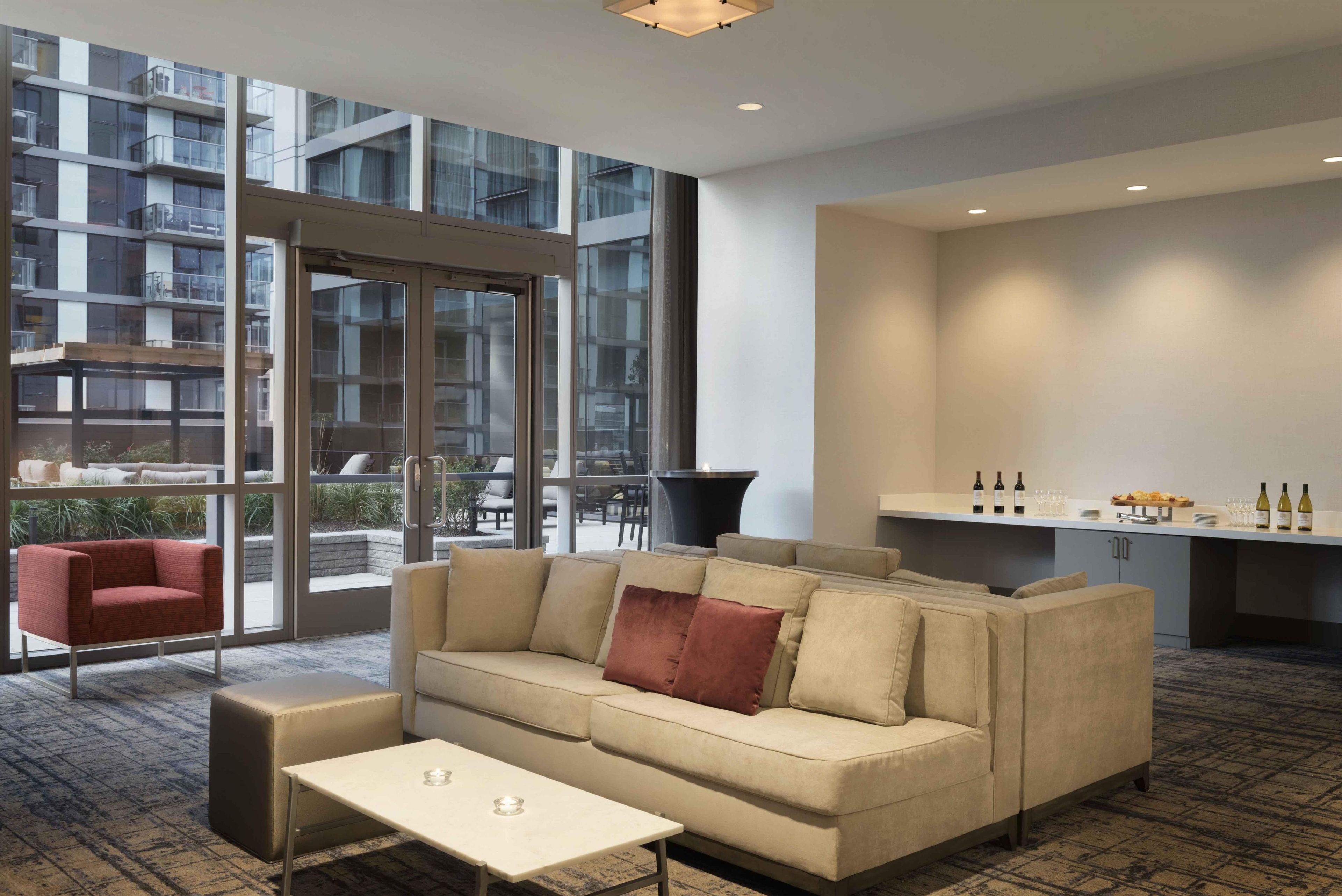 A modern hotel room with a sofa, a small table, a kitchen area with wine bottles, and large windows offering a view of the building's exterior.
