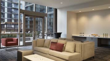 Ein modernes Hotelzimmer mit einer Couch, einem kleinen Tisch, einer Küchenzeile mit Weinflaschen und großen Fenstern mit Blick auf die Außenansicht des Gebäudes.