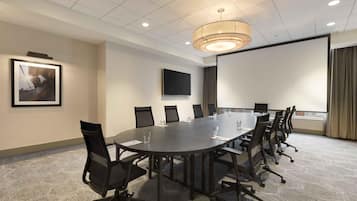 A conference room with a long table, chairs, a projector screen, and a television on the wall.