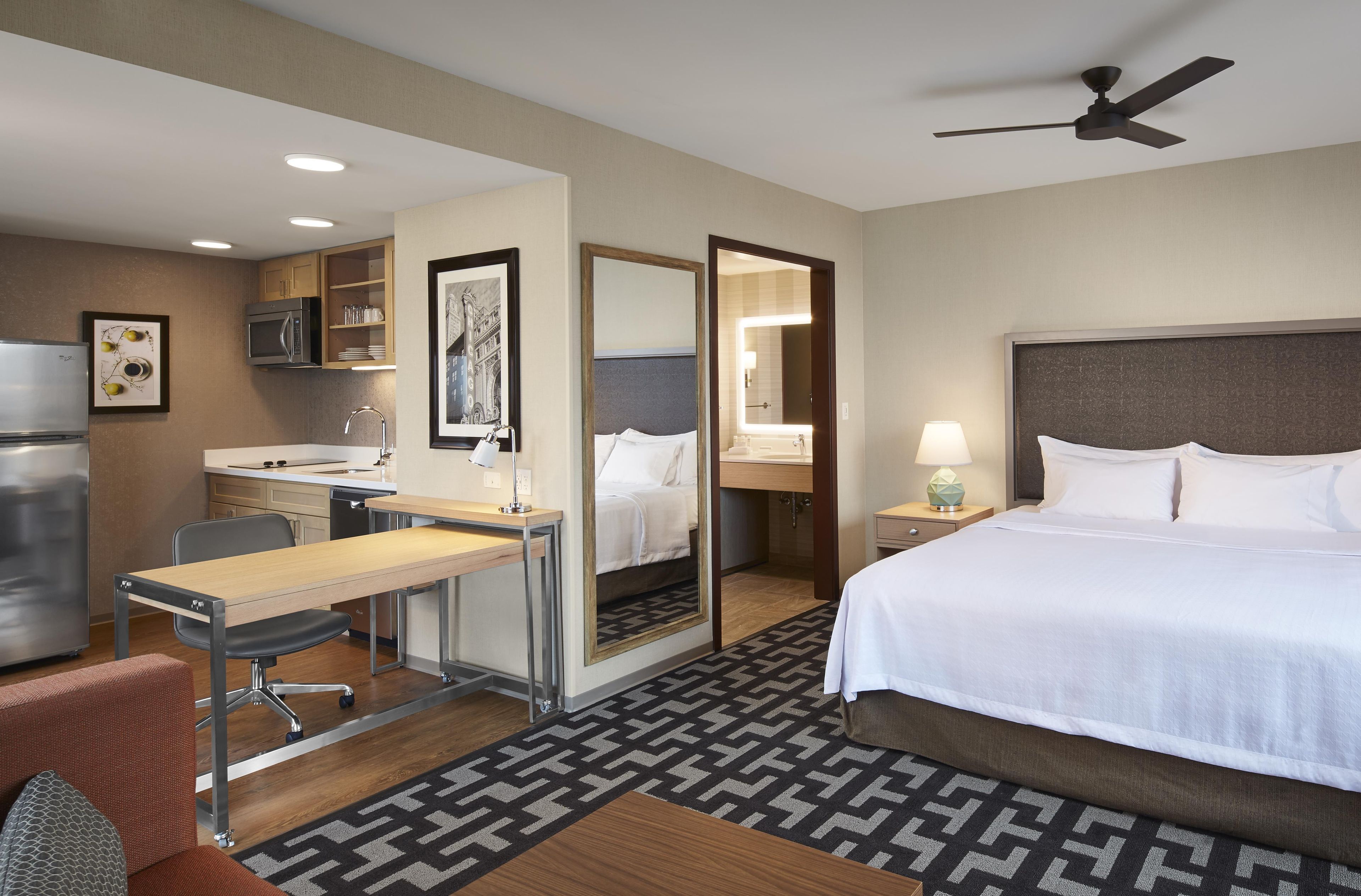 A hotel room with a bed, desk, chair, kitchenette, and a ceiling fan.