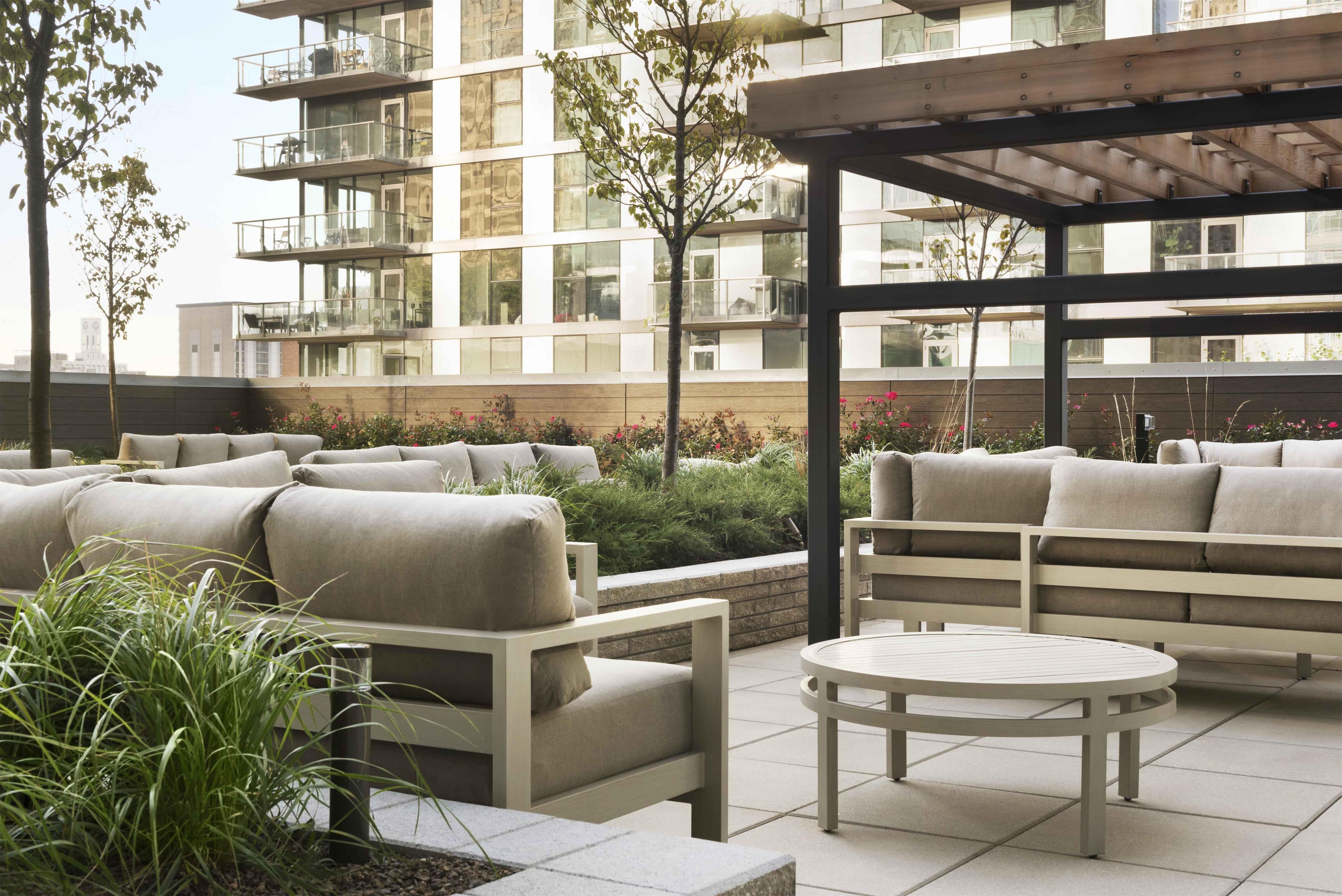 A modern outdoor seating area with sofas and a round table, surrounded by greenery and overlooking a multi-story building.