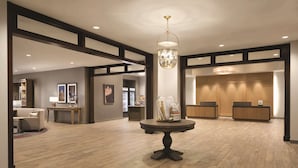 Un hall d’entrée spacieux doté d’un plancher en bois, une table ronde centrale agrémentée d’objets décoratifs, un comptoir de réception et des œuvres d’art encadrées qui ornent les murs.
