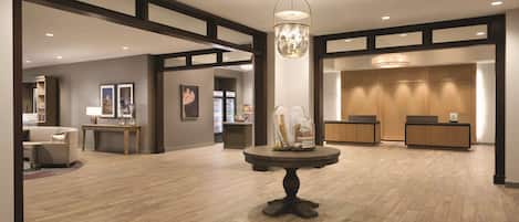 Un vestíbulo amplio con piso de madera, una mesa redonda central con elementos decorativos, un mostrador de recepción y obras de arte enmarcadas en las paredes.