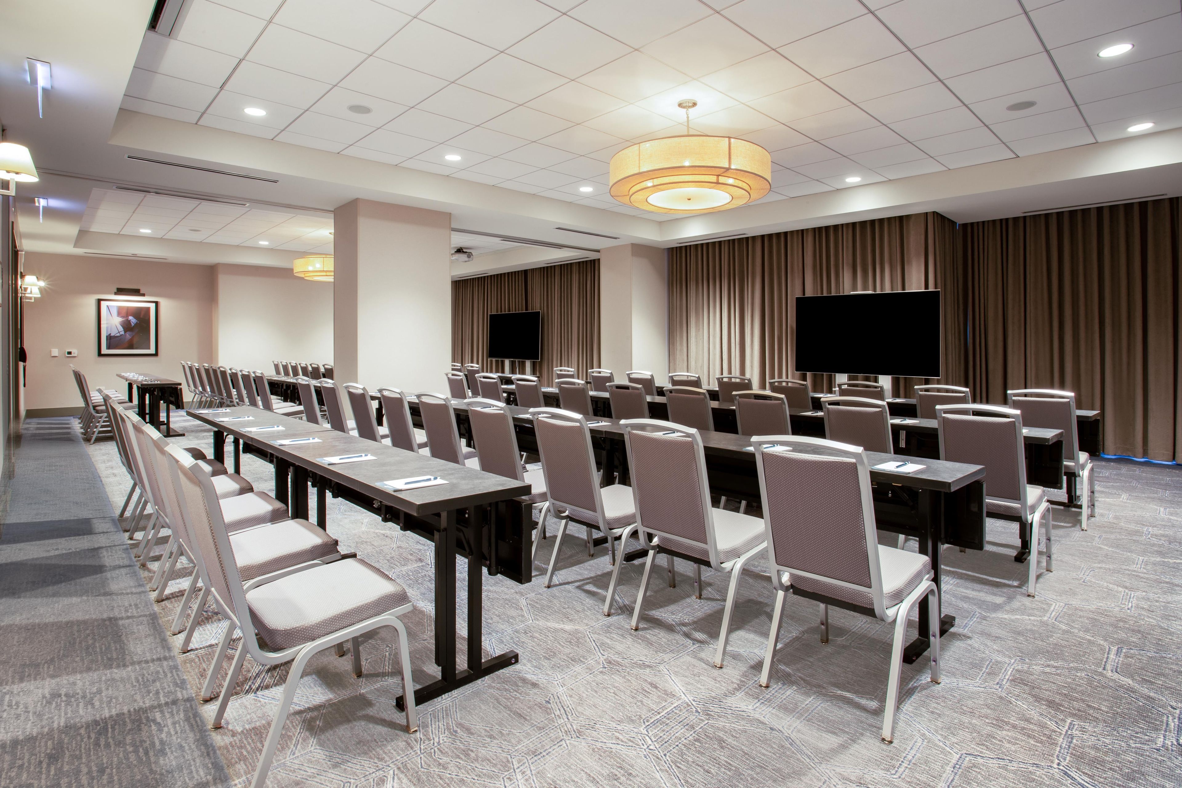 A conference room with rows of chairs, tables, a large screen, and a ceiling light fixture.