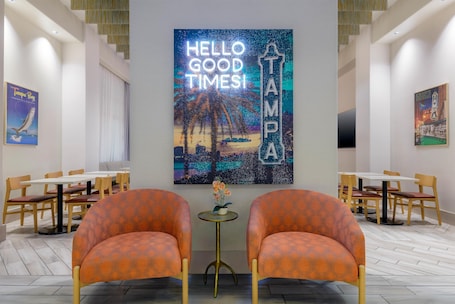 A hotel lobby with two orange armchairs, a 'Hello Good Times' sign, and a 'Tampa' sign. Tables and chairs are arranged around the room.. Hampton Inn & Suites Tampa Airport Avion Park Westshore