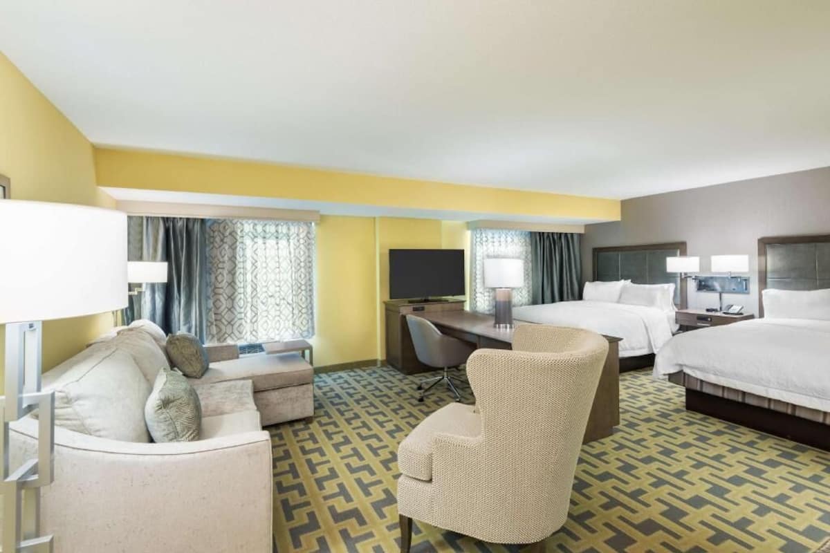 Une chambre d’hôtel équipée d’un canapé, d’un fauteuil, d’un bureau, d’une télévision, d’un lit et d’une fenêtre avec des rideaux.