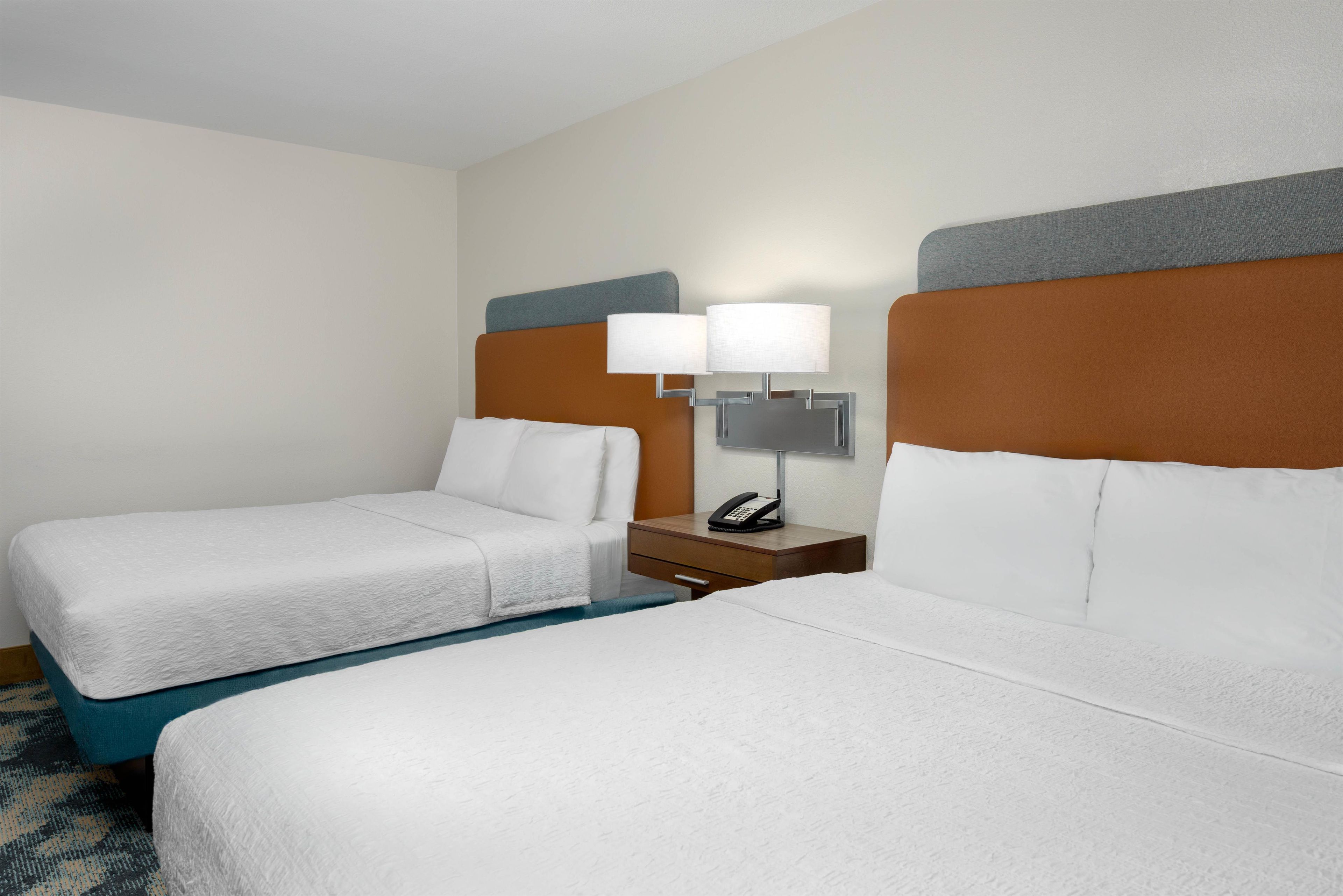 Habitación de hotel con dos camas, una mesita de noche con lámpara y teléfono, y piso alfombrado.