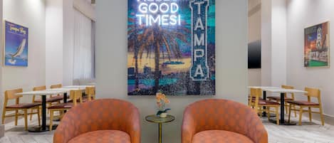 A hotel lobby with two orange patterned chairs, a large wall art piece with 'HELLO GOOD TIMES AMPA' text, and a poster of Tampa Bay.