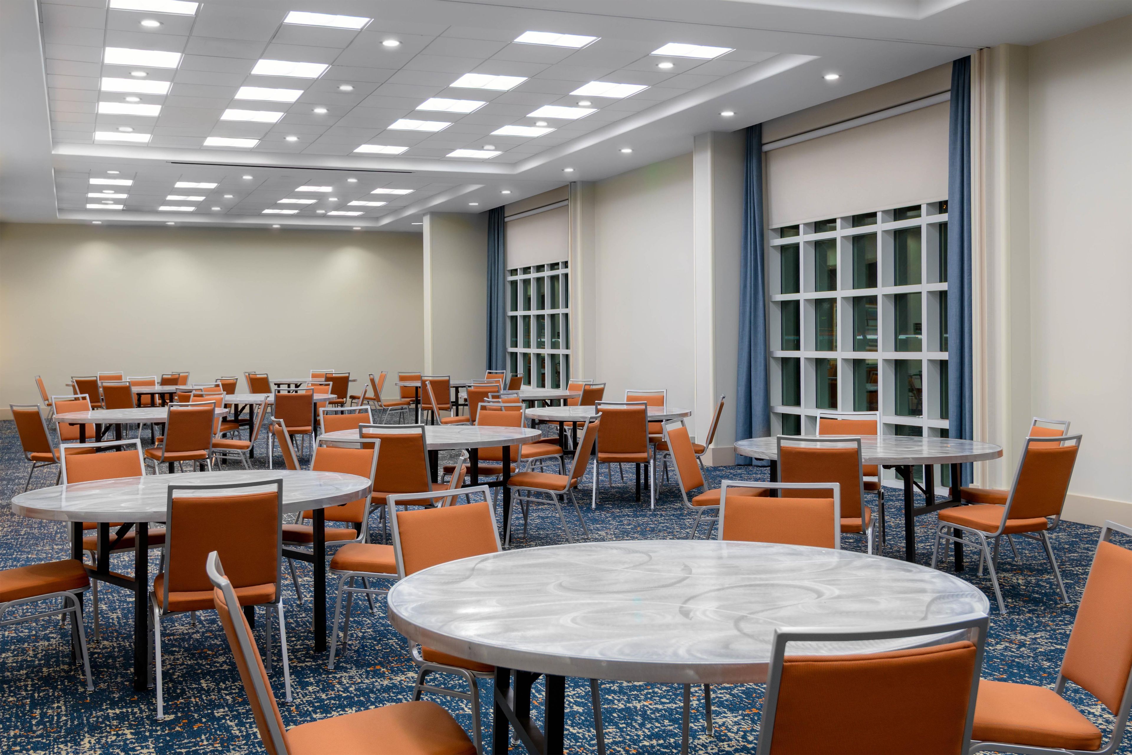 A conference room with multiple tables and chairs, large windows, and a carpeted floor.