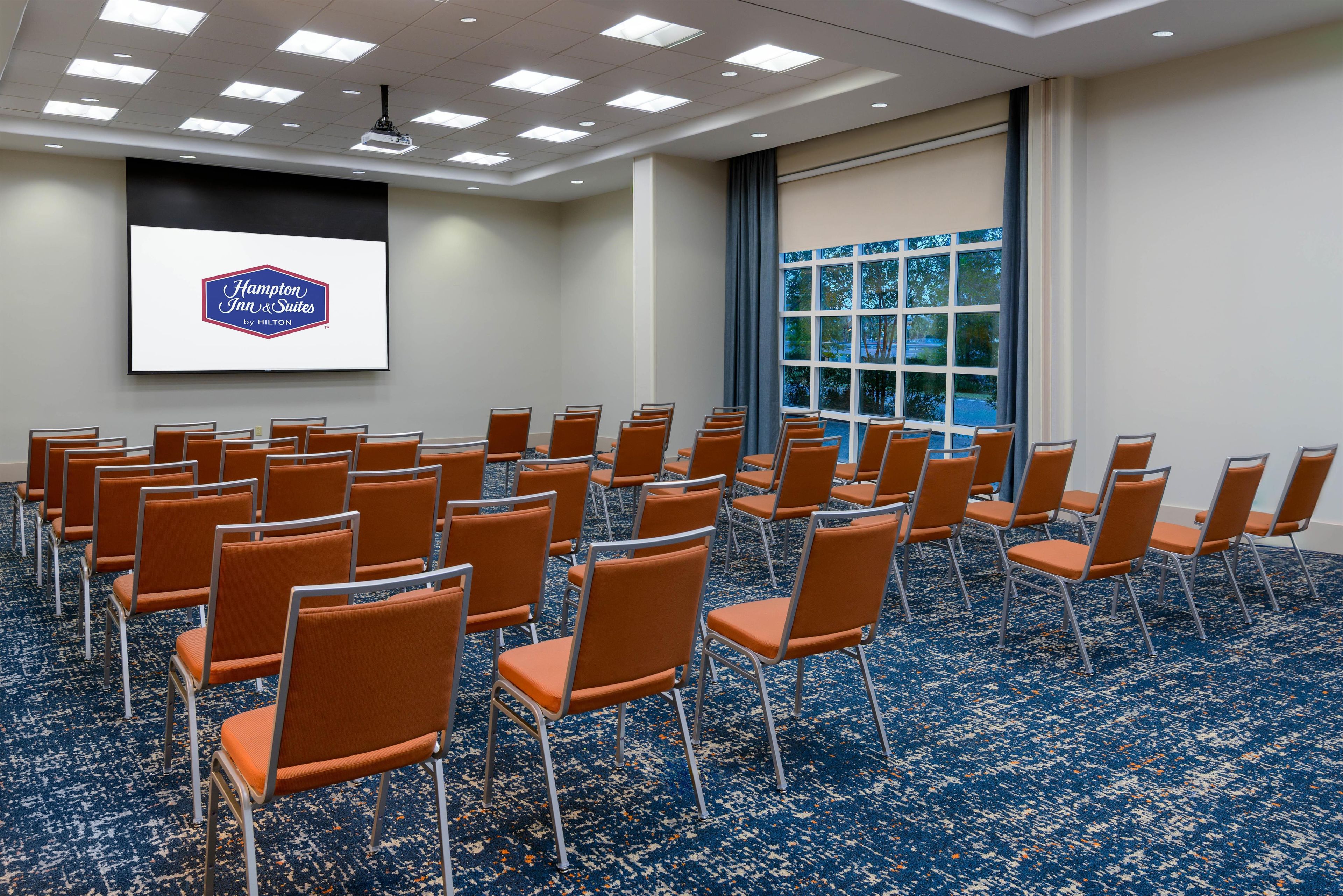 A conference room with rows of orange chairs, a large screen displaying a logo, and a large window with a view of trees.