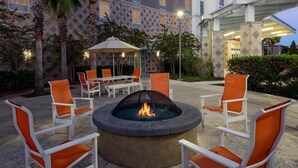 A fire pit surrounded by white chairs with orange cushions, set in an outdoor patio area with a patterned building in the background. - Hampton Inn & Suites Tampa Airport Avion Park Westshore (Tampa)
