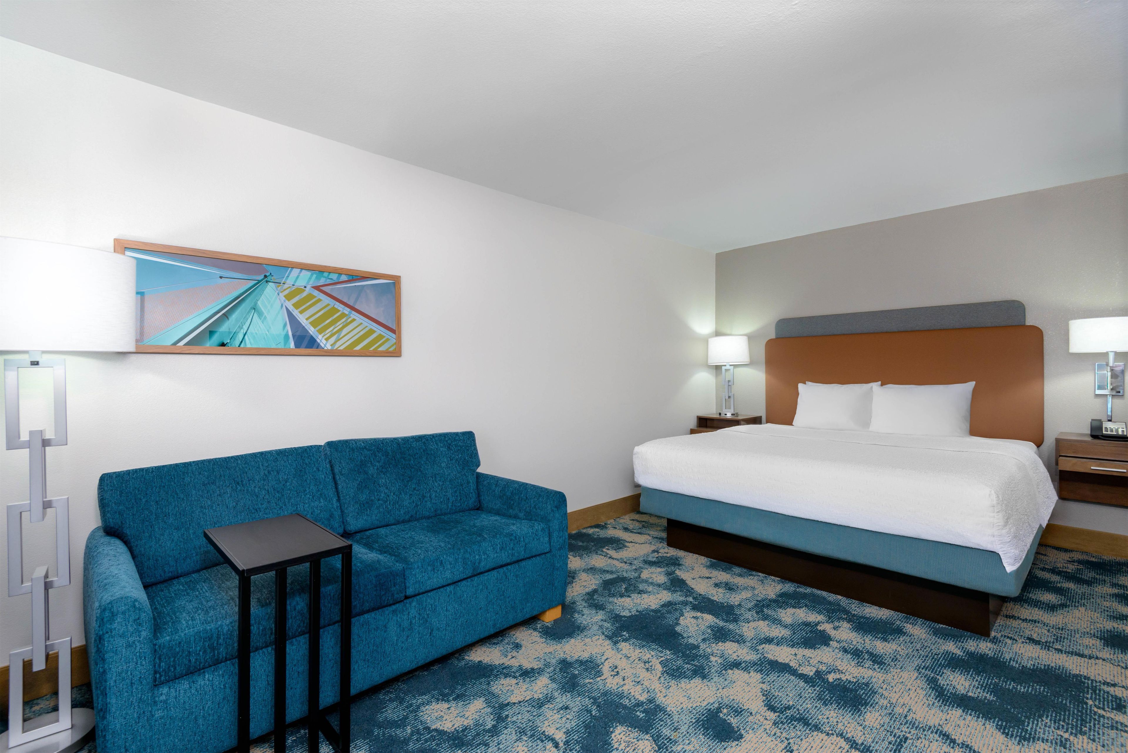 A hotel room with a bed, a blue sofa, a small table, a lamp, and a painting on the wall.