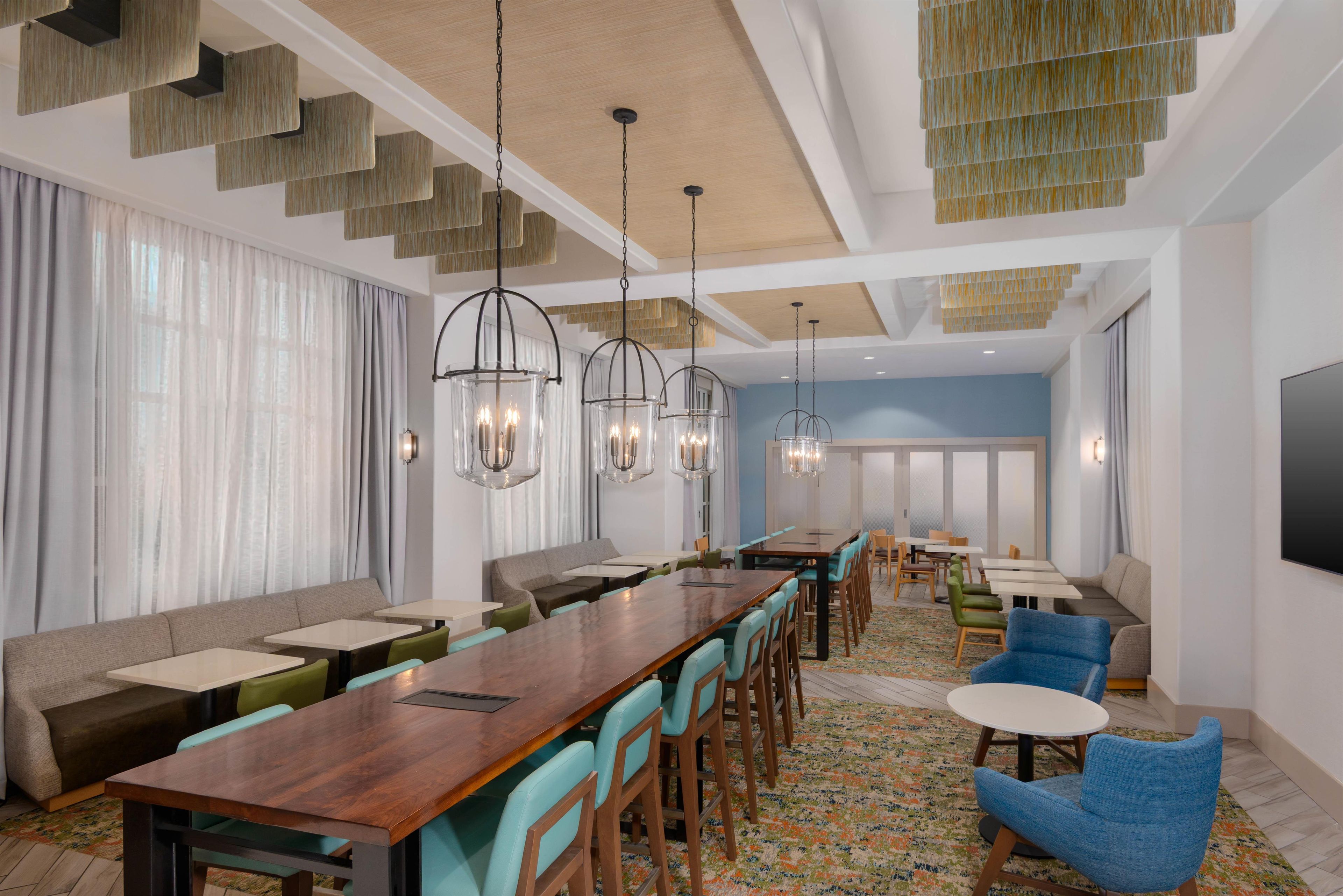 A modern dining area with a long wooden table, multiple chairs, and hanging pendant lights.