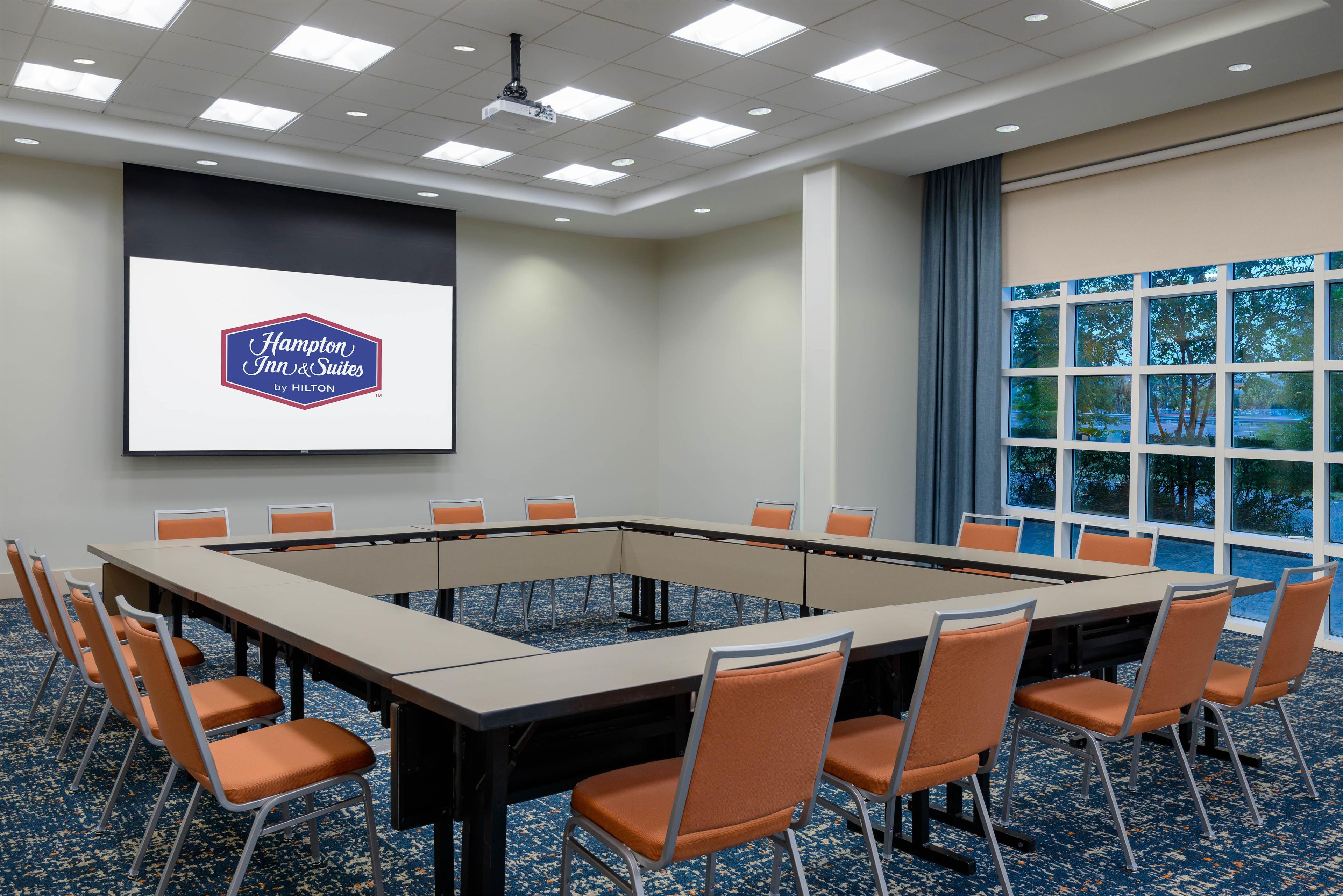 A conference room with a large table and chairs, a projector screen with the Hampton Inn & Suites logo, and a view of trees through large windows.