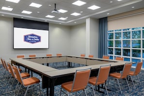 A conference room with a large table and chairs, a projector screen with the Hampton Inn & Suites logo, and a view of trees through large windows. - Hampton Inn & Suites Tampa Airport Avion Park Westshore (Tampa)