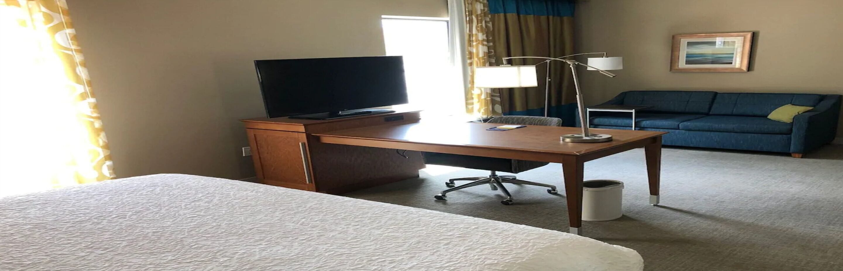 Ein Hotelzimmer mit einem Bett, einem Schreibtisch, einem Flachbildfernseher, einer Lampe, einem blauen Sofa und einem Bild an der Wand.