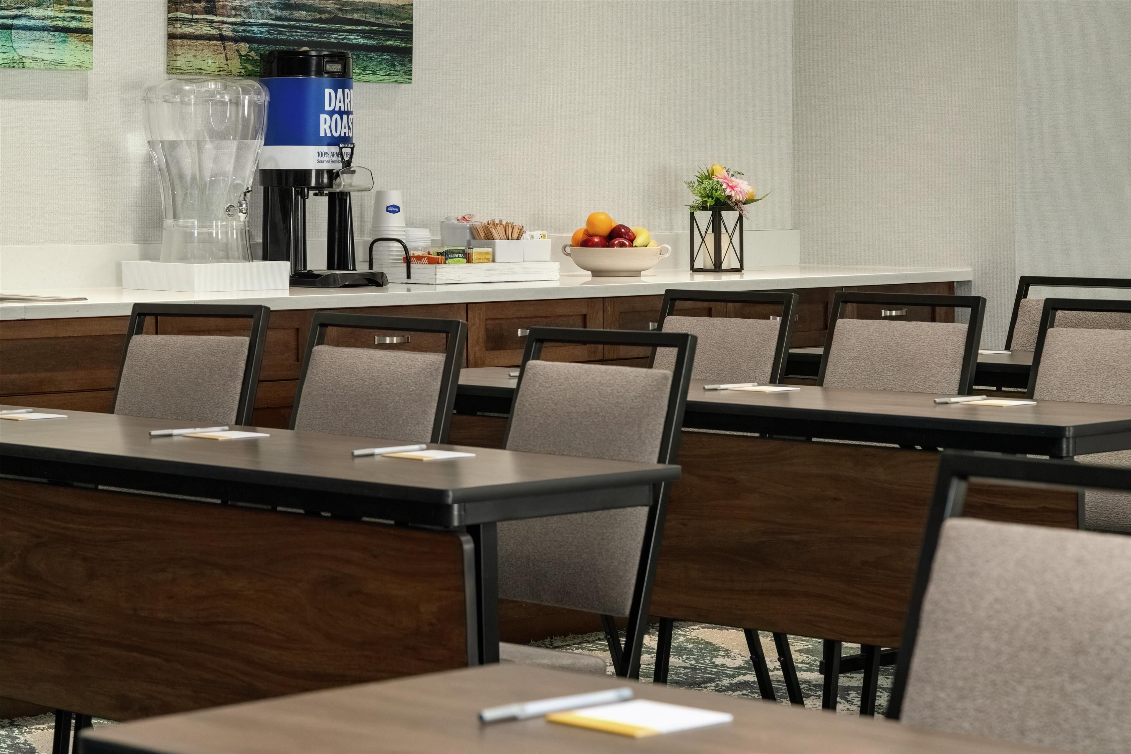 Uma sala de conferências com várias mesas e cadeiras, uma máquina de café, uma cesta de frutas e um vaso com flores.