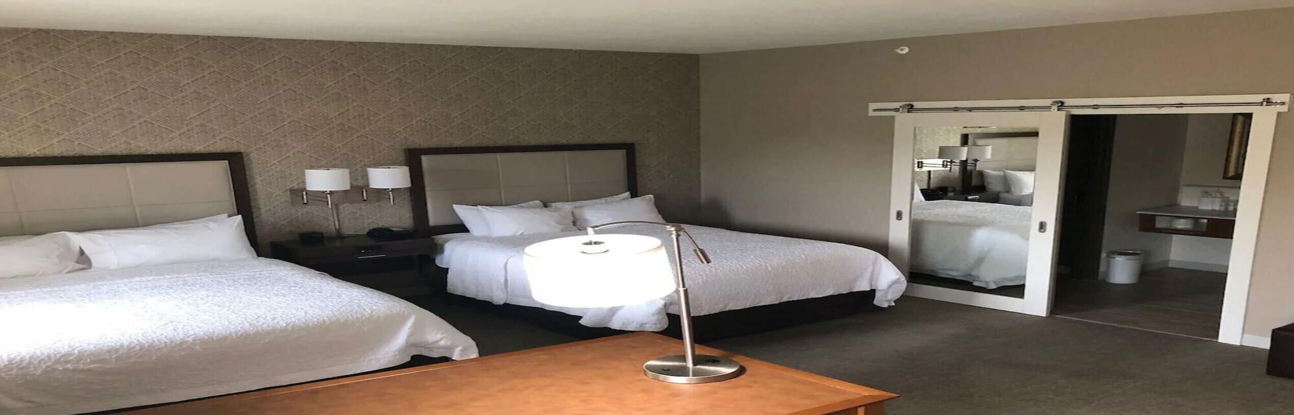 A hotel room with two beds, a bedside table, a lamp, a mirror, and a sliding door to a bathroom.