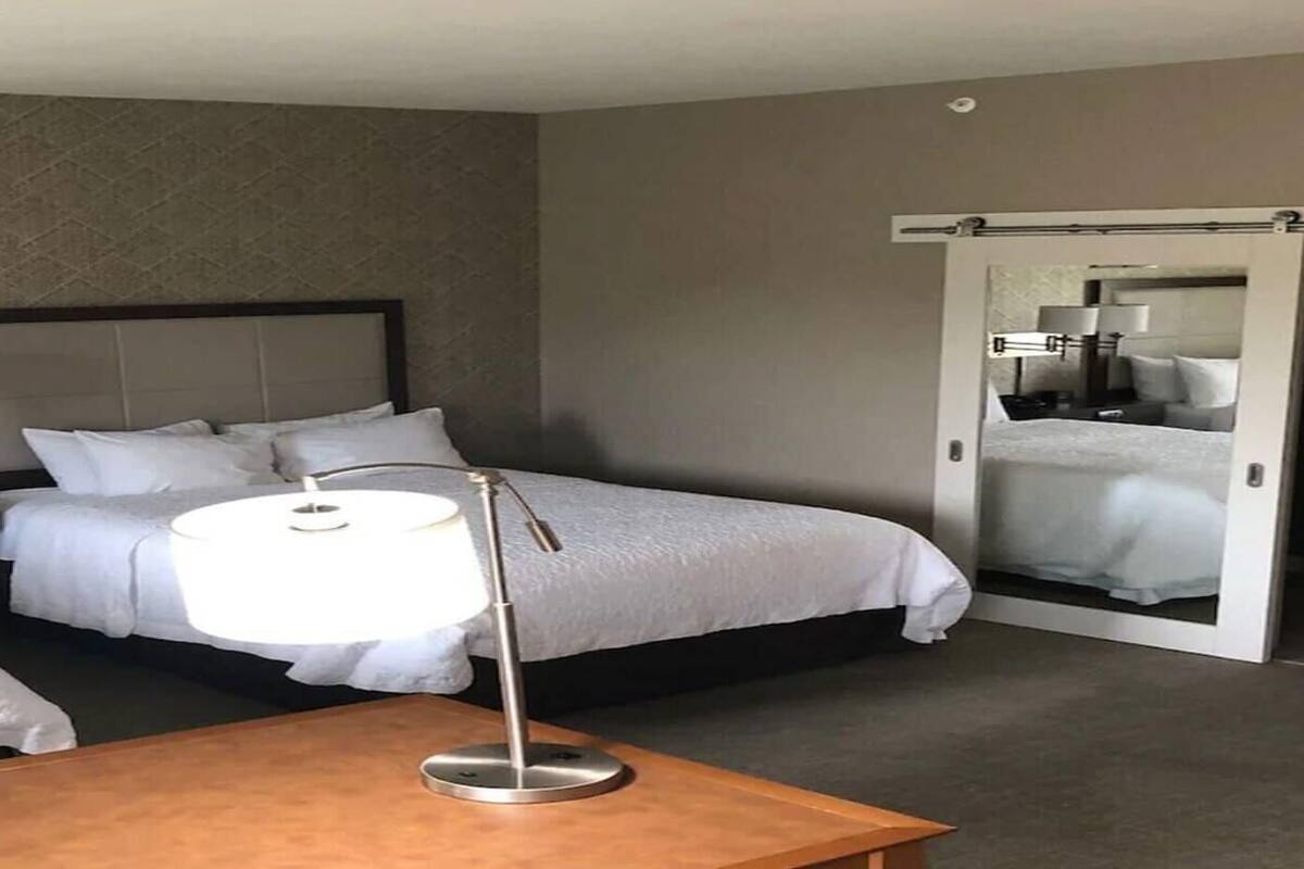 A hotel room with two beds, a bedside table, a lamp, a mirror, and a sliding door to a bathroom.