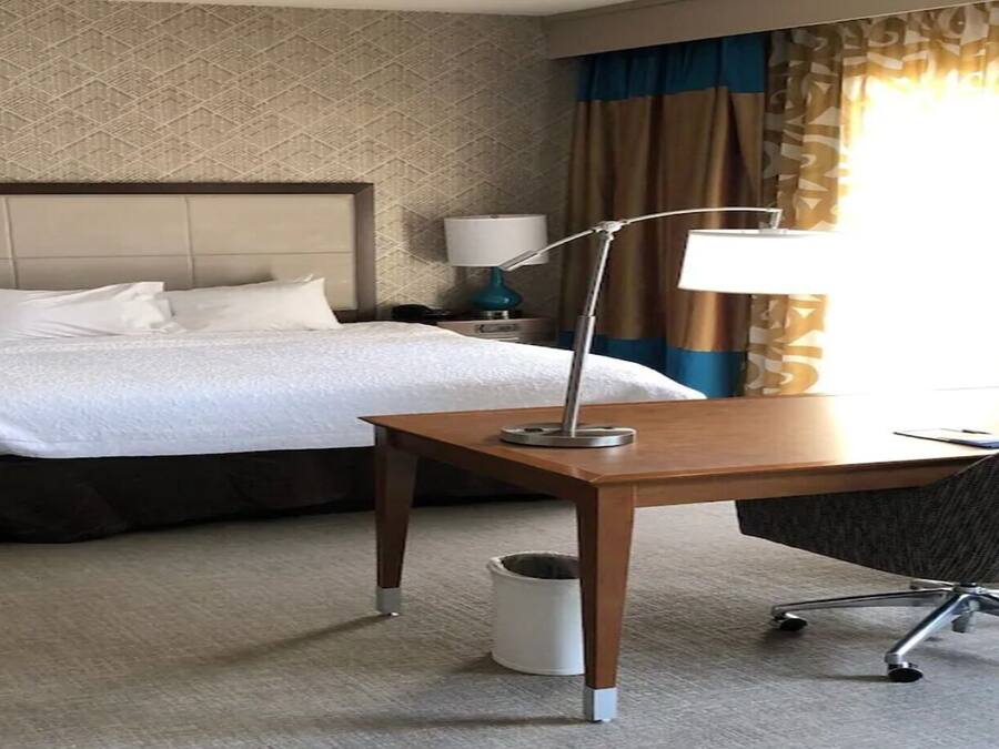 Quarto de hotel com cama, escrivaninha, cadeira, televisão, luminárias e cortinas.