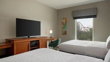 Habitación de hotel con dos camas, un televisor sobre un escritorio de madera, una lámpara de escritorio, una silla y una ventana con vistas a una montaña rusa.