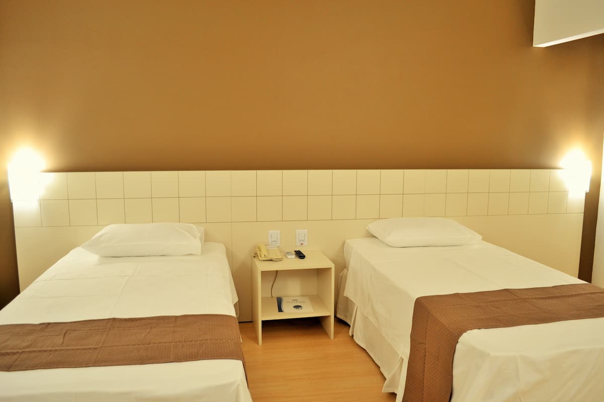 Una habitación de hotel con dos camas individuales, una mesita de noche con un teléfono y un control remoto, y una lámpara fijada a la pared.