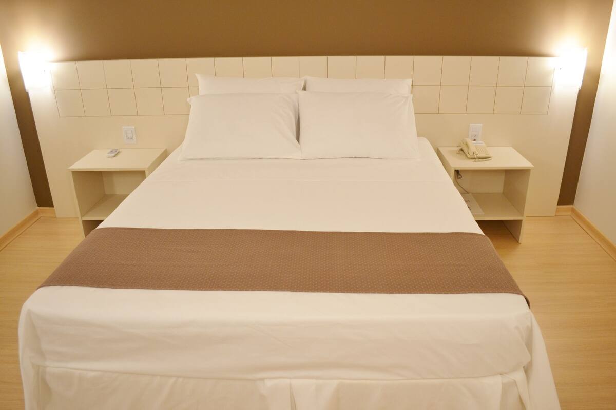 Una habitación de hotel con una cama grande, dos mesitas de noche y un cabecero con una lámpara.