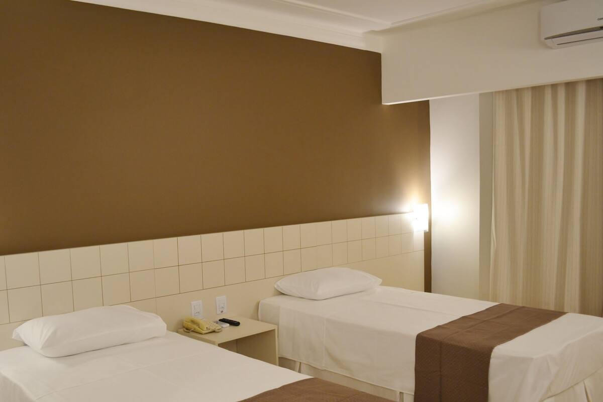 Habitación de hotel con dos camas, una mesita de noche con teléfono y control remoto, aire acondicionado de pared y ventana con cortinas.