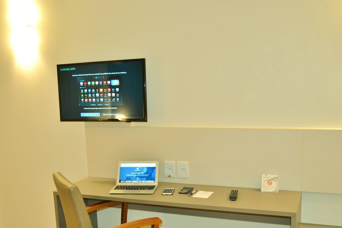 Una habitación de hotel con escritorio, computadora portátil, silla, televisión montada en la pared y algunos objetos sobre el escritorio, como un teléfono, un control remoto y un bloc de notas.