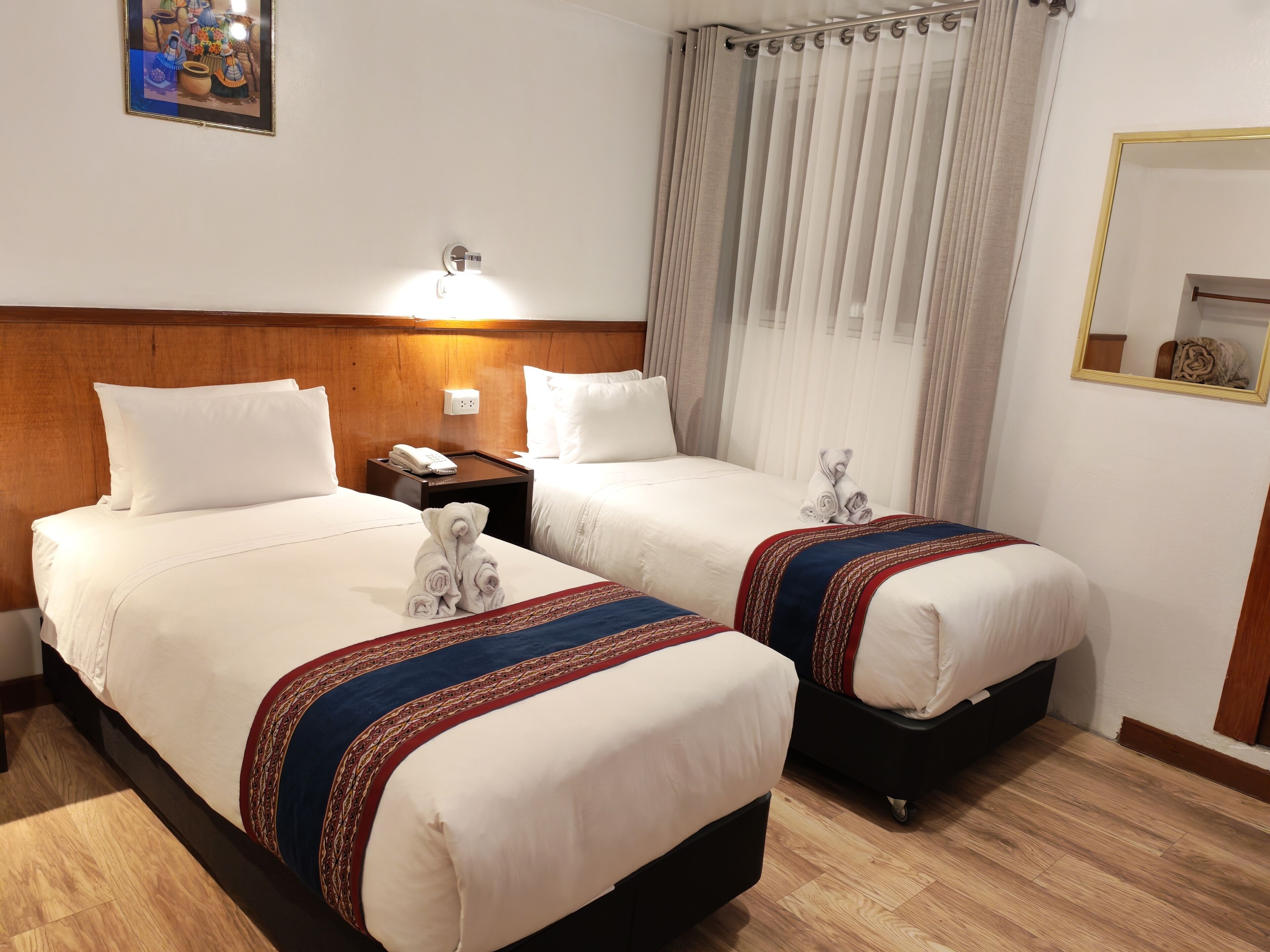 Habitación de hotel con dos camas, una mesita de noche con teléfono, una lámpara de pared, una ventana con cortinas y un espejo.