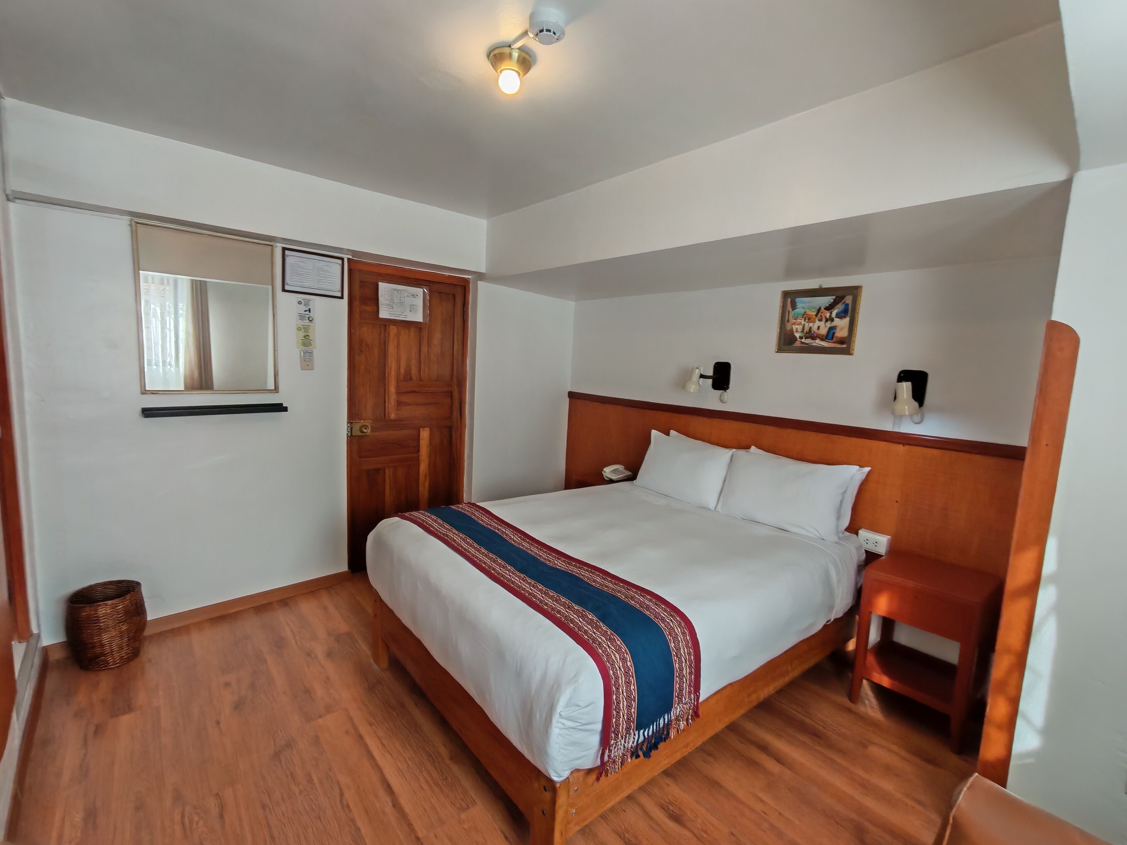 A hotel room with a wooden bed, a wooden door, a small wooden stool, a wall-mounted telephone, and a ceiling light fixture.