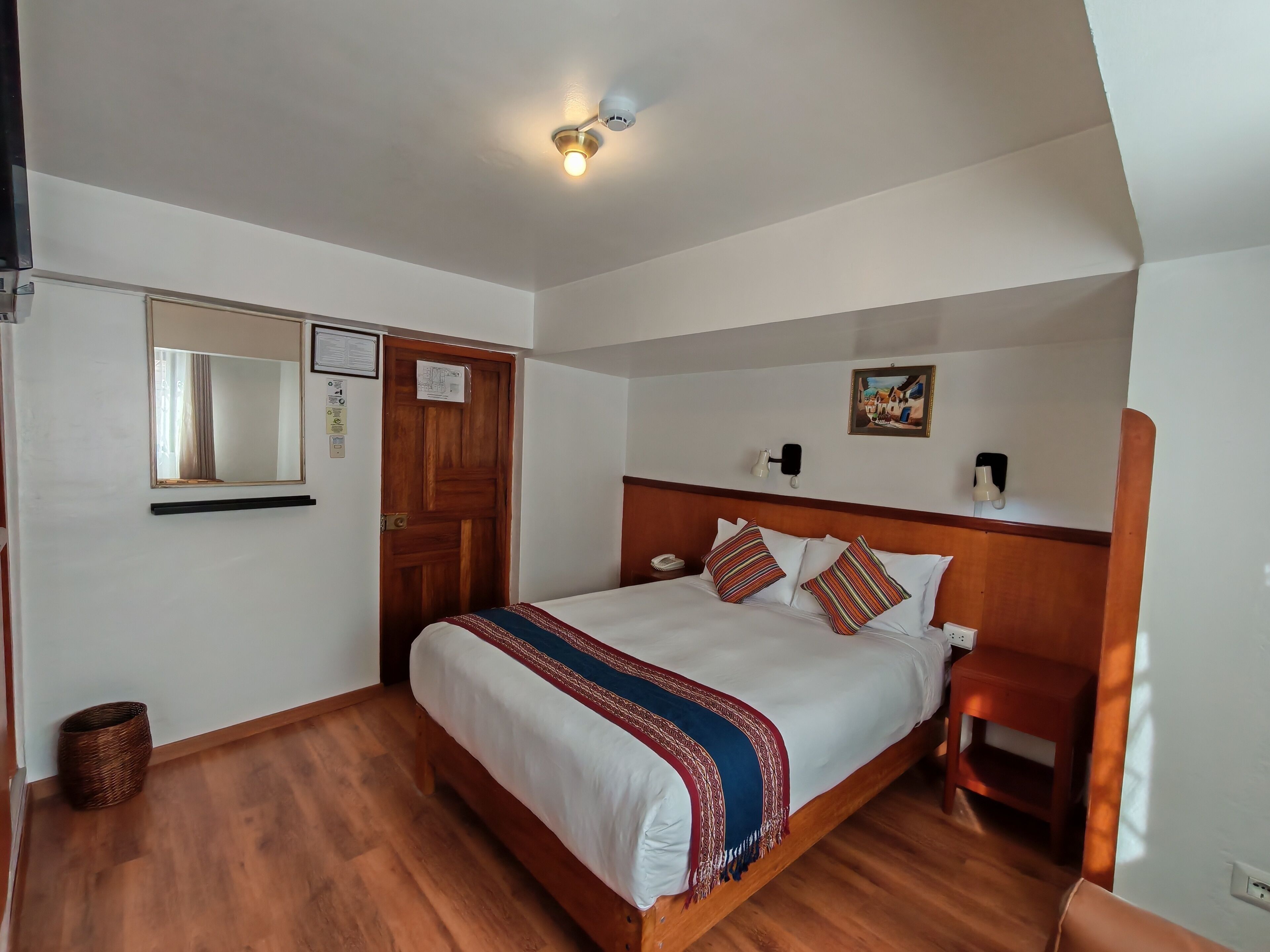 A hotel room with a wooden bed, a nightstand, a wall-mounted lamp, a ceiling light, a wooden door, and a mirror.