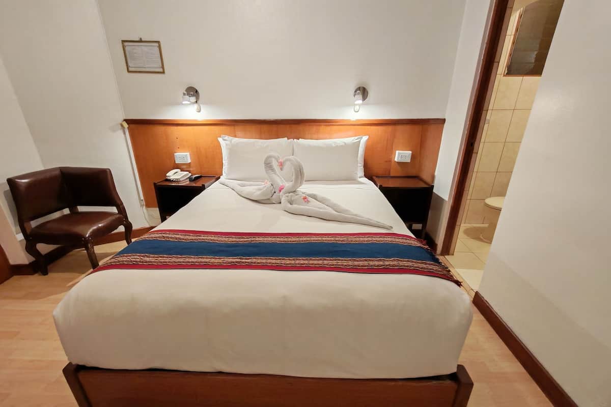 A hotel room with a wooden headboard, a bed with white linens, a decorative swan towel, a brown leather chair, and a nightstand with a phone and lamp.